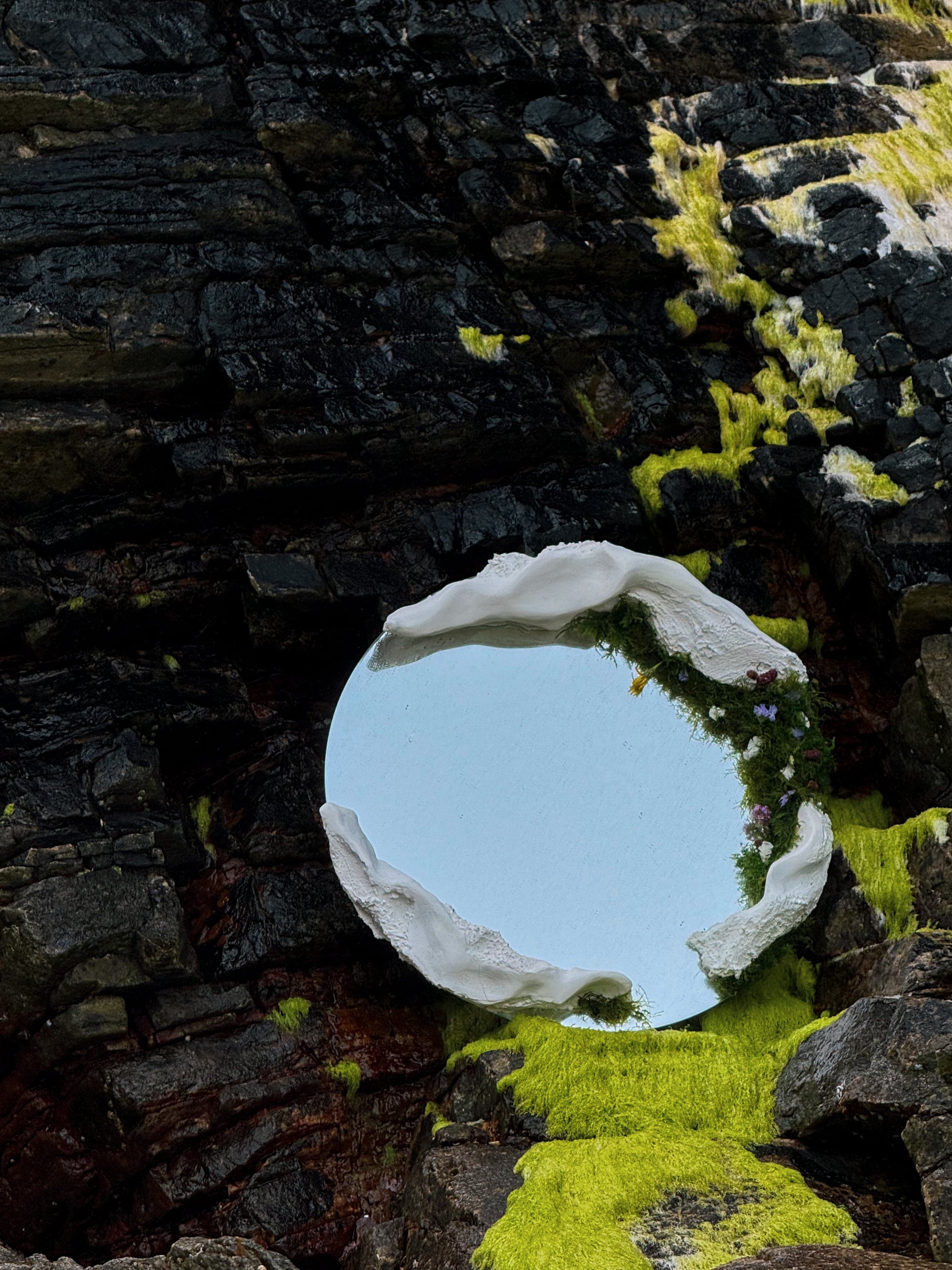 Large round mirror held by white sculptural fragments and moss against dark wet rock
